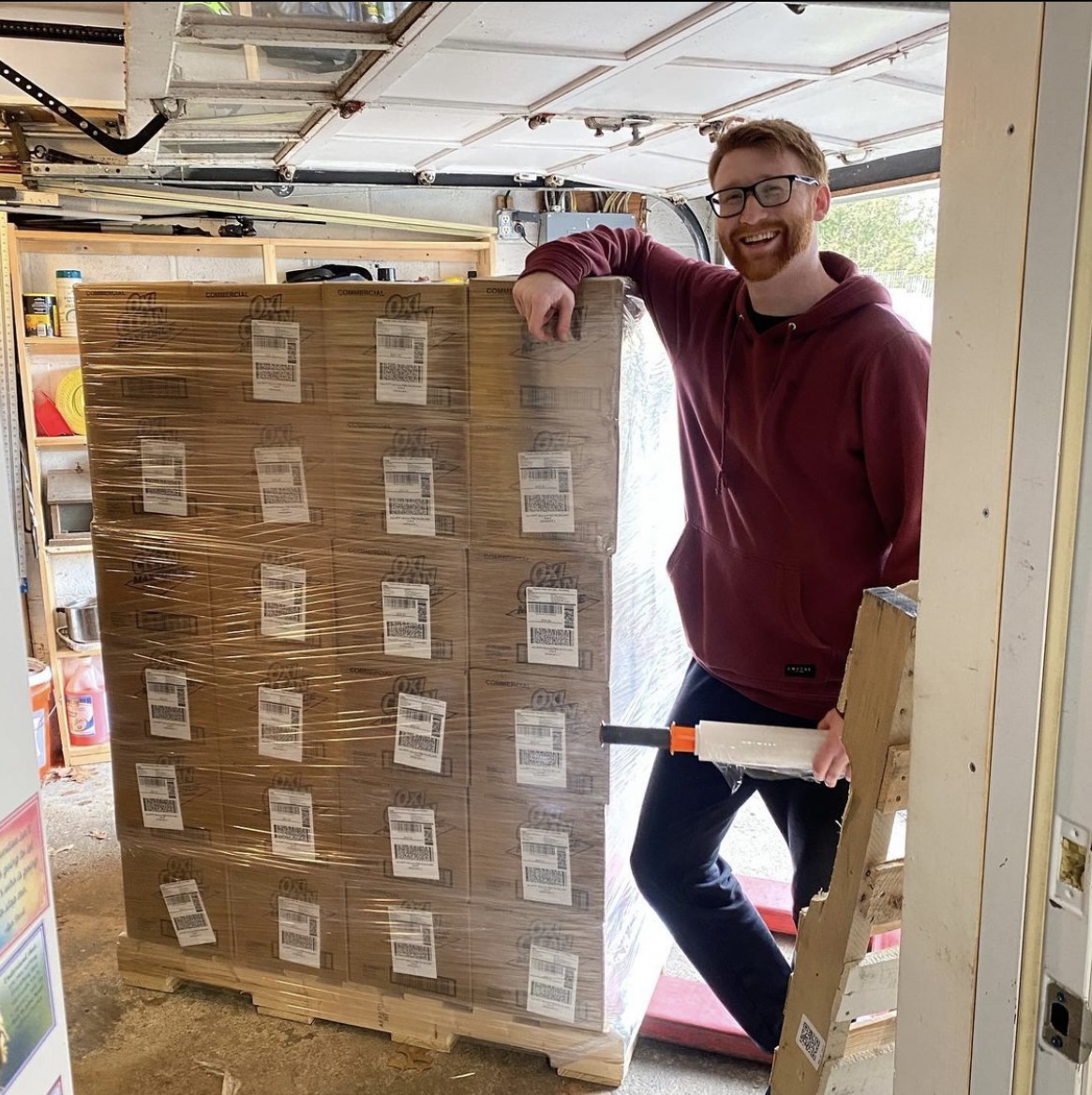 The founder in his warehouse, standing beside a wrapped pallet of inventory and holding a stretch-wrap roll
